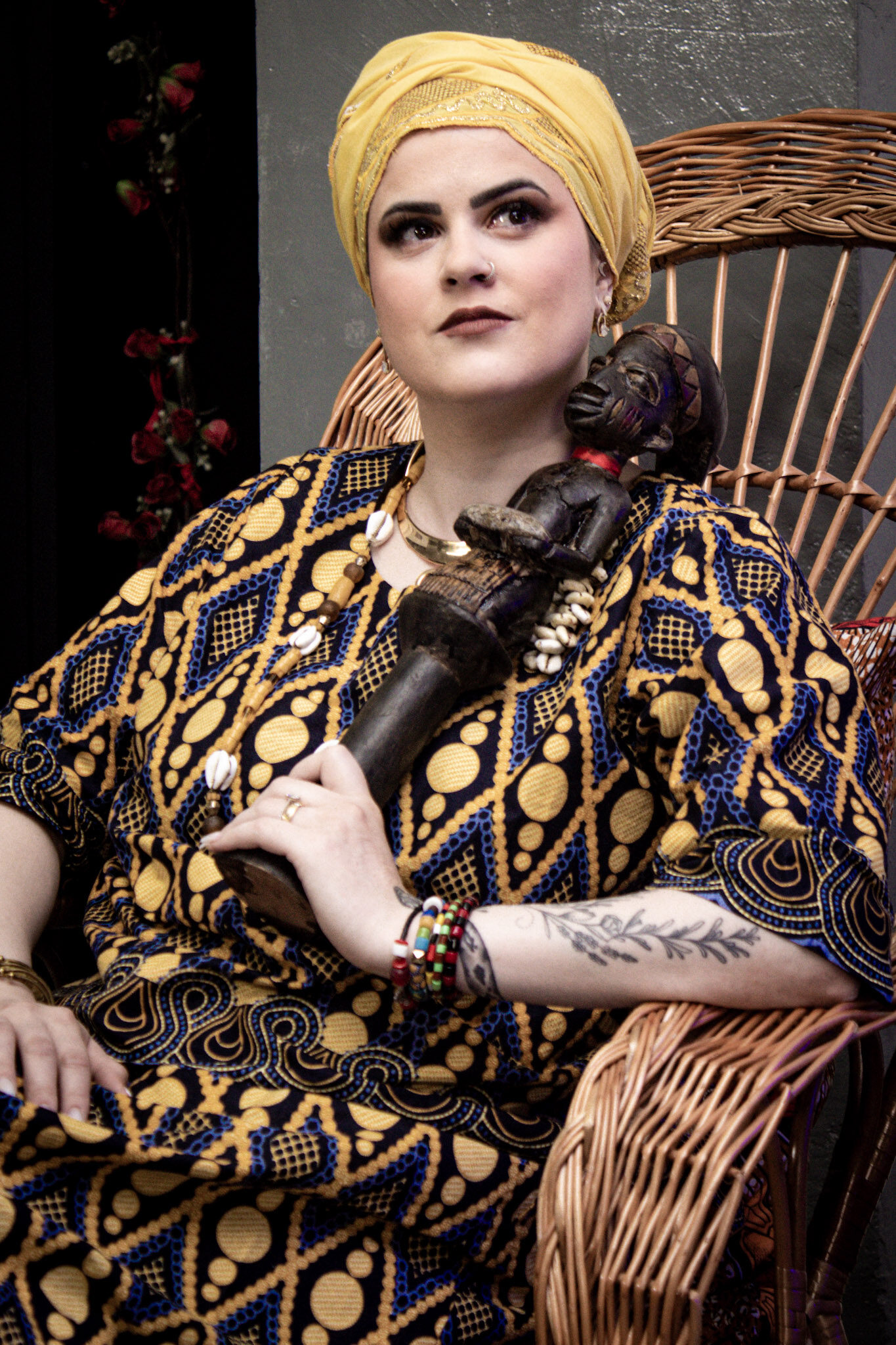 Mãe Taciana, Ìyálórìṣà Fádamilolà, sentada em cadeira de vime com gele amarelo e agbádá de estampa azul e dourada, segurando o ọ̀ṣẹ́ de Ṣàngó